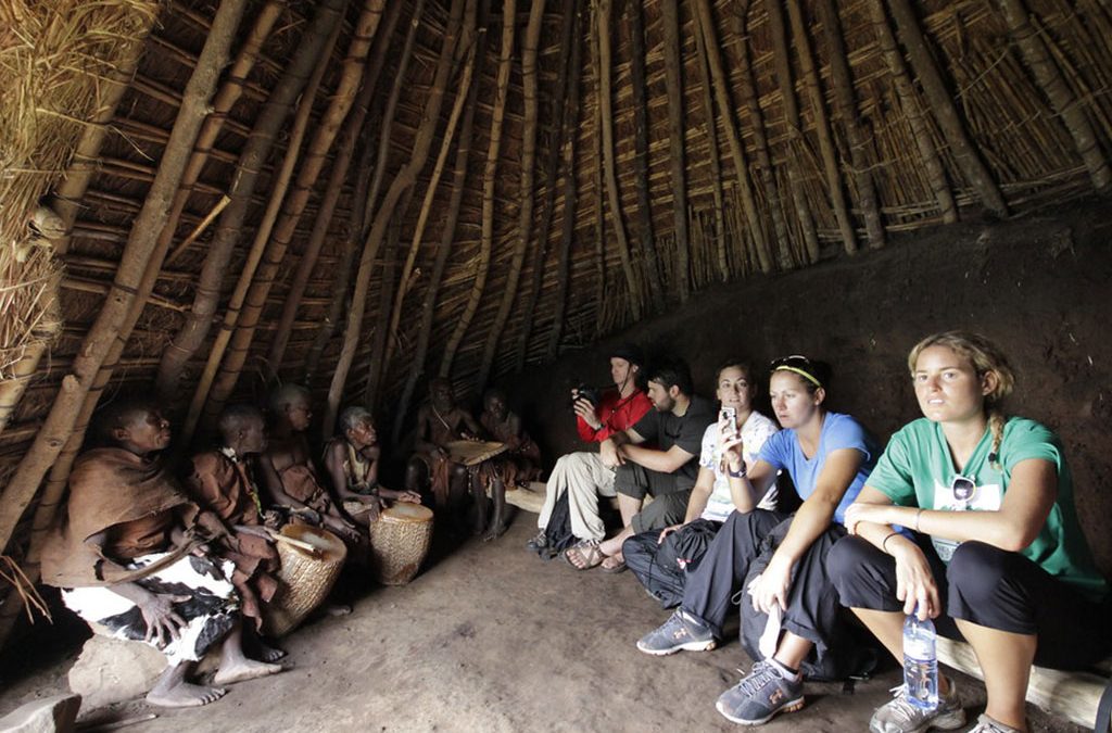 batwa-hut-gathering-with-tourists - Mumwe Safaris Uganda Group of travelers sitting along the right side of a thatched hut, watching villagers drum on the left interior wall.