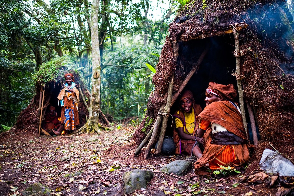 batwas-of-uganda - Mumwe Safaris Uganda People in traditional clothing sit near a rustic mud-and-wood hut in a dense forest clearing.