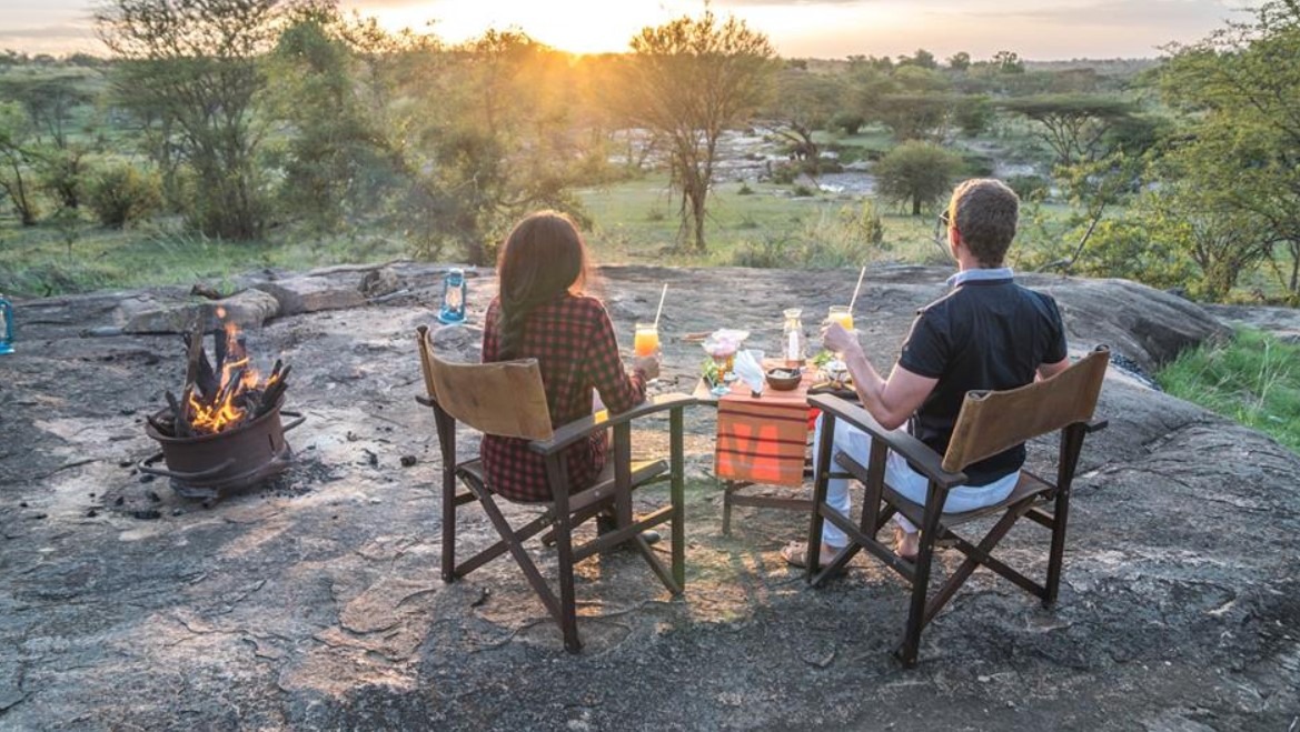 Couple seated in wooden chairs at a rocky overlook during sunset, sharing a meal with a campfire nearby.