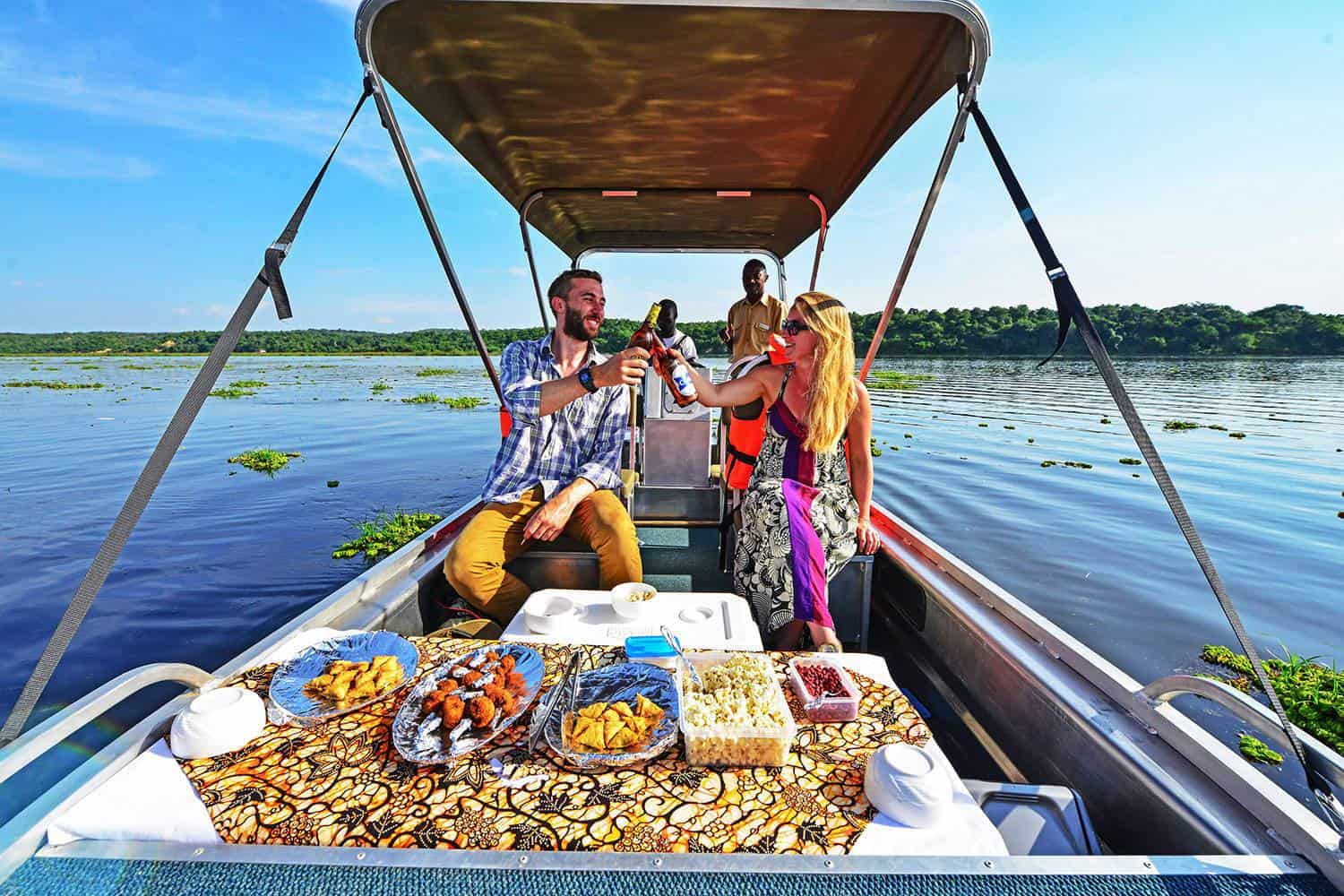 Two people on a boat share a toast over a table of snacks as water and greenery pass by in the background.
