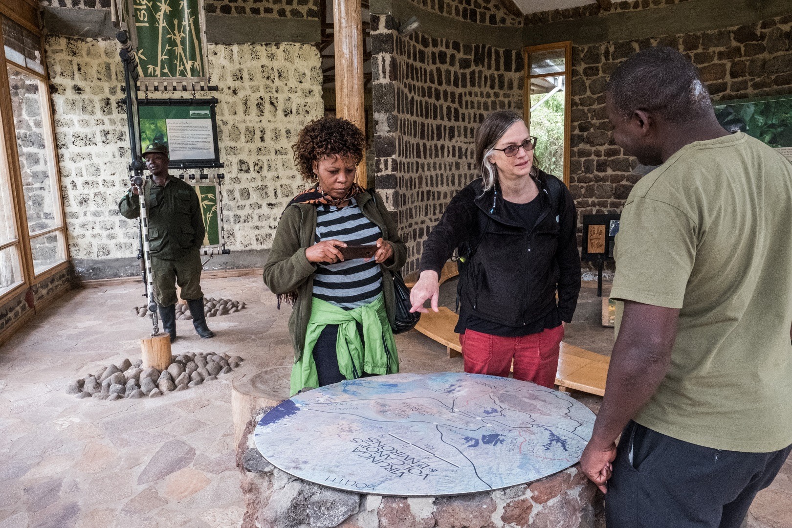 - Mumwe Safaris Uganda Visitors study a large circular map on a stone pedestal inside a rustic museum; a guide in green uniform stands nearby.