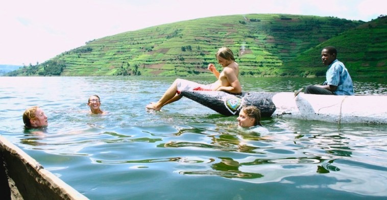 Group of children swimming in a calm lake while a person sits on a small boat and watches, with terraced green hills in the background.